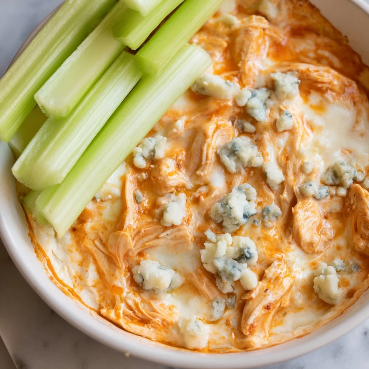 A close-up of golden-brown Spicy Buffalo Chicken Dip in a dish, with bright green celery sticks ready for serving.