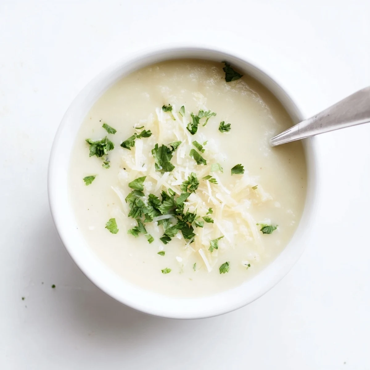 A bowl of steaming Asiago Roasted Garlic Cauliflower Soup is garnished with fresh chives and extra grated cheese beside a slice of crusty bread.